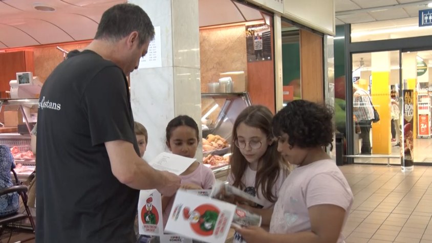 Els més petits ensenyen la llista de la compra a un dels comerciants del mercat cobert